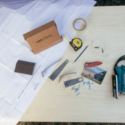 Laid out contents of the vancillary sprinter headliner shelf DIY kit on a sheet of plywood showing the paper template, mounting brackets, hardware, as well as the required tools, jigsaw, pencil, measuring tape, which is all you need to build a DIY sprinter van headliner shelf.