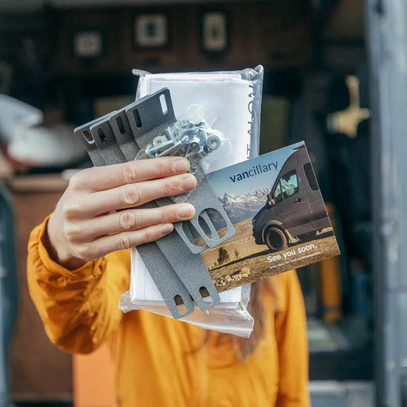 woman holding sprinter diy conversion kit, brackets, paper template and mounting hardware standing in front of a mercedes sprinter van.