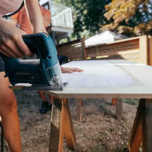 Woman cutting plywood with jigsaw for sprinter van headliner shelf using paper template taped onto plywood.